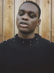 Portrait of a Black man in his 20s wearing a black ribbed sweater with silver studs against a wooden fence.