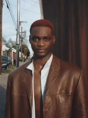 Portrait of a Black man with red hair wearing a brown leather jacket and unbuttoned white shirt against an outdoor street backdrop.