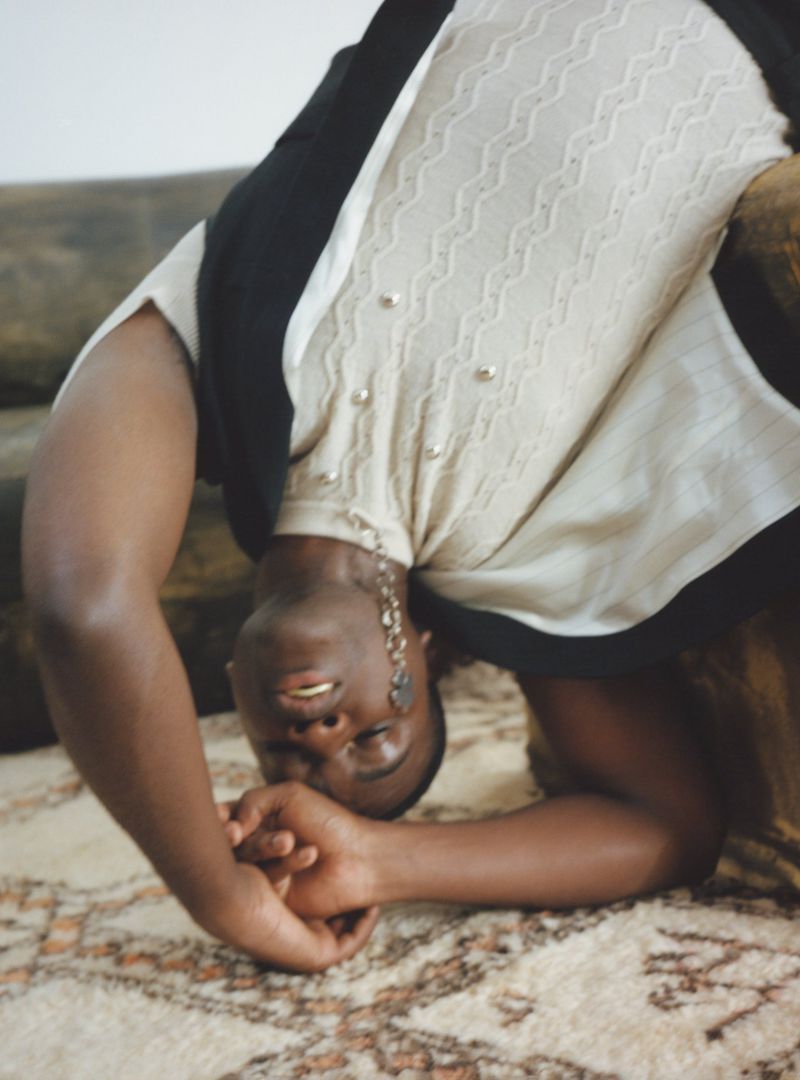 Model arching backward wearing a textured cream knit top and dark vest on a patterned rug.