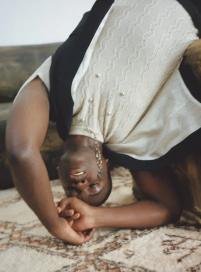 Model arching backward wearing a textured cream knit top and dark vest on a patterned rug.