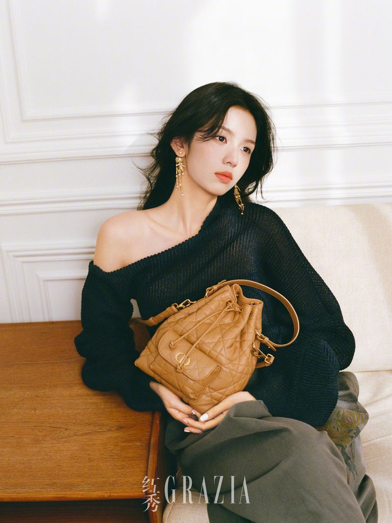 A young woman with long dark hair poses indoors next to a wooden table with a brown leather drawstring bag in her lap.