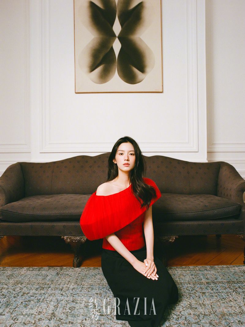 A seated young Asian woman with long dark hair, wearing a red asymmetrical top and black skirt, poses indoors in front of a brown couch.