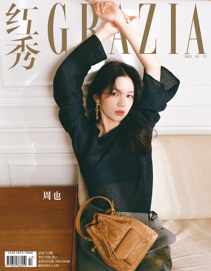 Magazine cover shot of an adult female model with dark hair posing with her arms raised above her head and holding a tan purse.