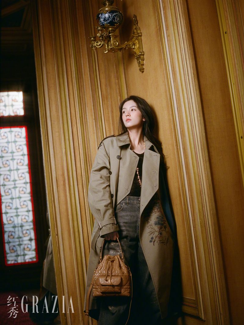 A woman in a tan trench coat poses indoors with a brown handbag. She leans against a wall with decorative moulding and an ornate wall lamp.