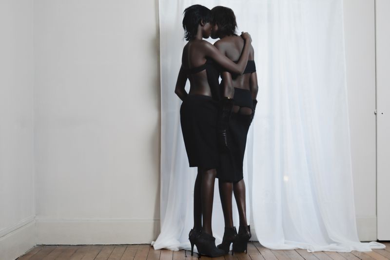 Two dark-skinned women in black outfits embrace closely in front of a white wall and sheer curtains, standing on a hardwood floor.