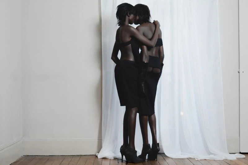 Two dark-skinned women in black outfits embrace closely in front of a white wall and sheer curtains, standing on a hardwood floor.