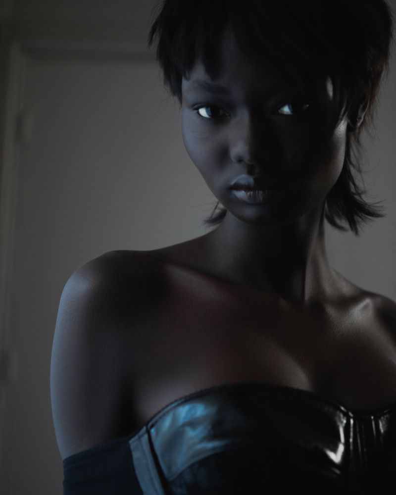 Portrait of a young woman with short dark hair and a glossy black strapless top, gazing forward in a dimly lit indoor setting.
