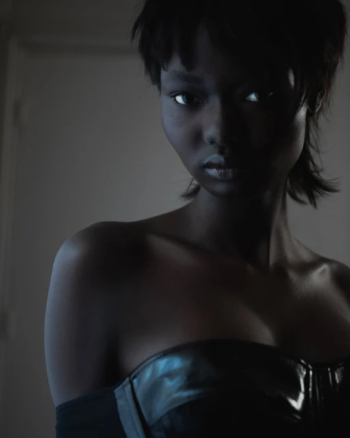 Portrait of a young woman with short dark hair and a glossy black strapless top, gazing forward in a dimly lit indoor setting.