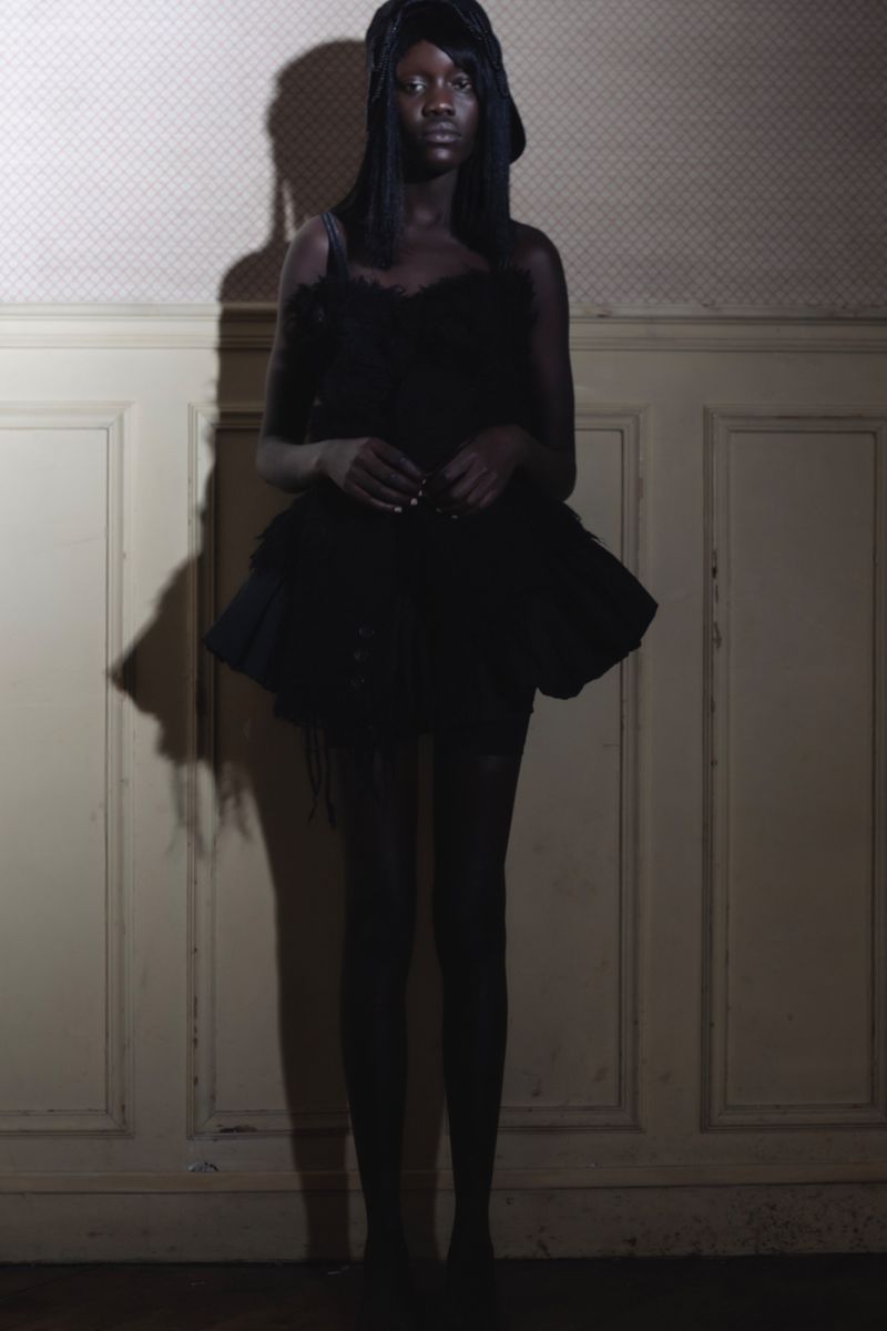Full-length studio shot of a Black female fashion model in a dark textured dress and head covering, posing against a muted wall.