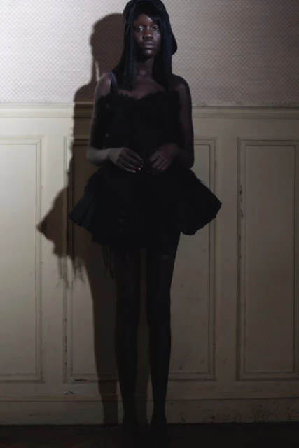 Full-length studio shot of a Black female fashion model in a dark textured dress and head covering, posing against a muted wall.