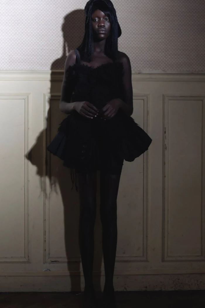 Black woman wearing a dark ruffled dress standing against a paneled wall with a prominent shadow cast behind her.