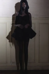 Black woman wearing a dark ruffled dress standing against a paneled wall with a prominent shadow cast behind her.