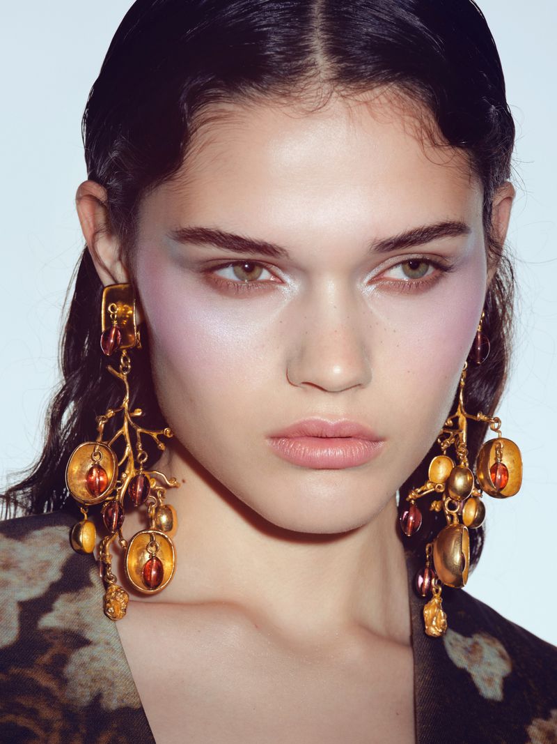 Close-up of a woman wearing ornate gold earrings, her face adorned with dramatic lavender blush highlighting her cheekbones.