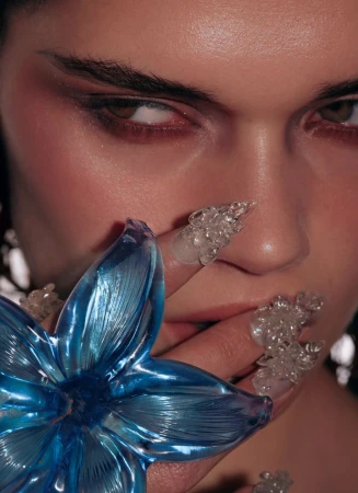 Close-up of a woman's face with creative acrylic nail art and a blue flower. Dramatic lighting highlights makeup and nail details.