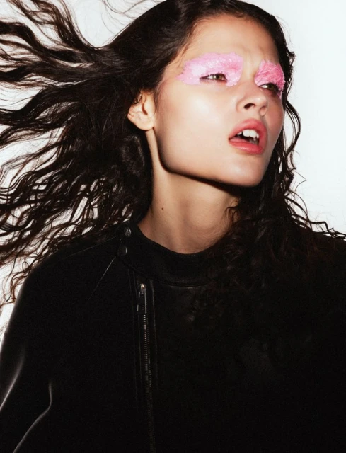 Young woman with wind-blown dark hair, leather jacket, pink eye makeup poses against a white backdrop in a beauty fashion shot.