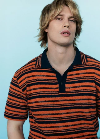 Close-up portrait of a blond male model wearing a striped orange and navy polo shirt against a light blue studio background.