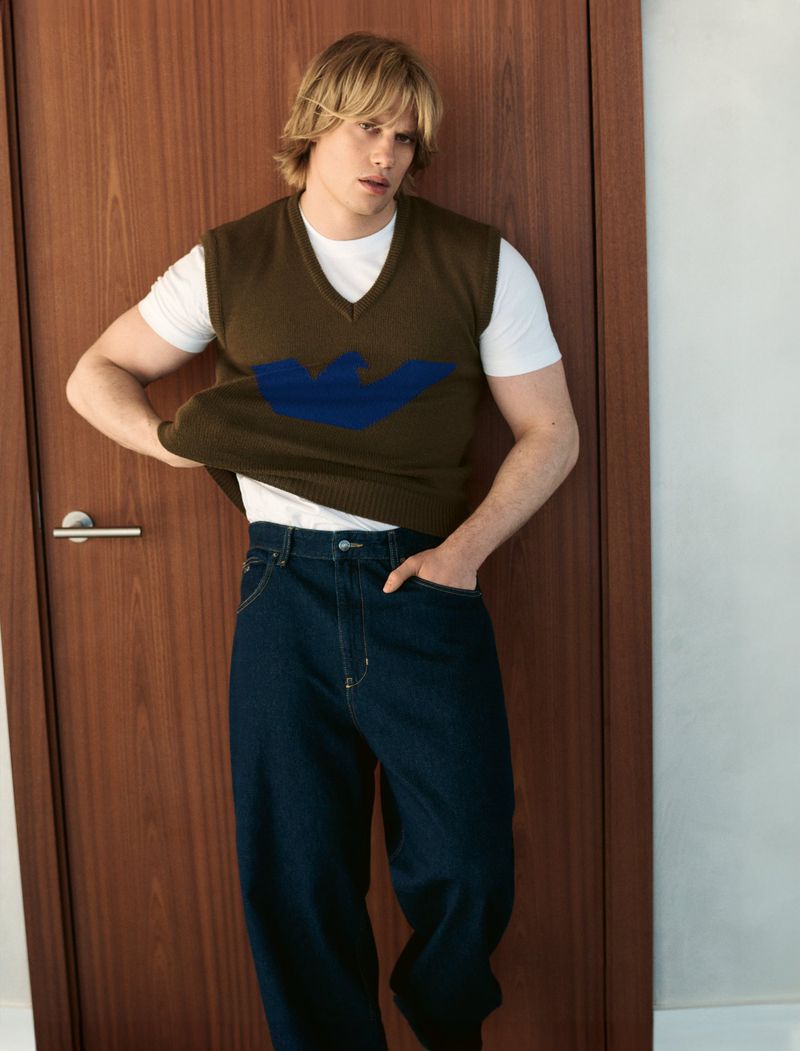 Blond male model poses indoors wearing a vest with a blue logo over a white t-shirt and blue jeans in a fashion setting.