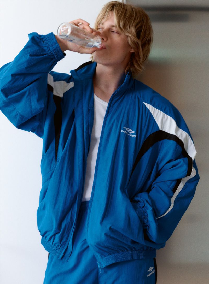 Young adult drinks water from a clear bottle wearing a blue Balenciaga tracksuit in a medium studio shot with neutral background.