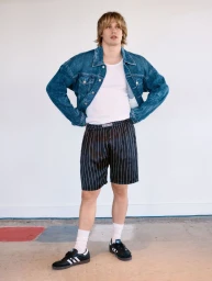Portrait of Nicholas Galitzine wearing a blue denim jacket, white tank top, and striped shorts against a plain background