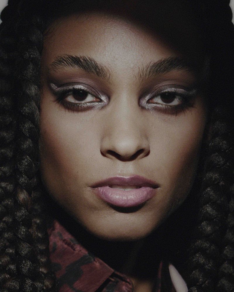 Close-up of an adult female model with dark hair and skin, wearing silver makeup and a patterned shirt in a dimly lit studio.
