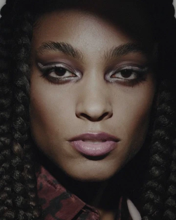 Close-up of an adult female model with dark hair and skin, wearing silver makeup and a patterned shirt in a dimly lit studio.