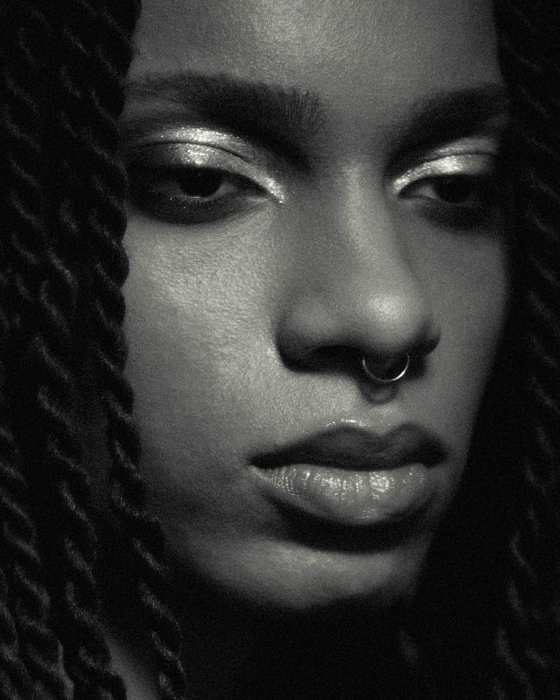 Close-up portrait of a Black woman with dark braided hair, silver eyeshadow, and a nose ring, showcasing beauty and makeup in a studio setting.