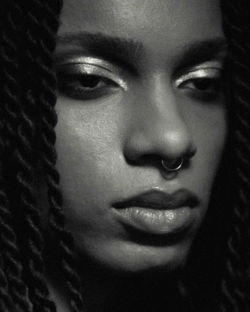 Close-up portrait of a Black woman with dark braided hair, silver eyeshadow, and a nose ring, showcasing beauty and makeup in a studio setting.
