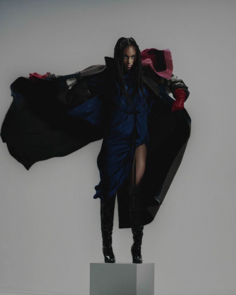 Fashionable woman poses in blue dress, black cape, and knee-high boots against a white backdrop on a gray platform, studio shot.