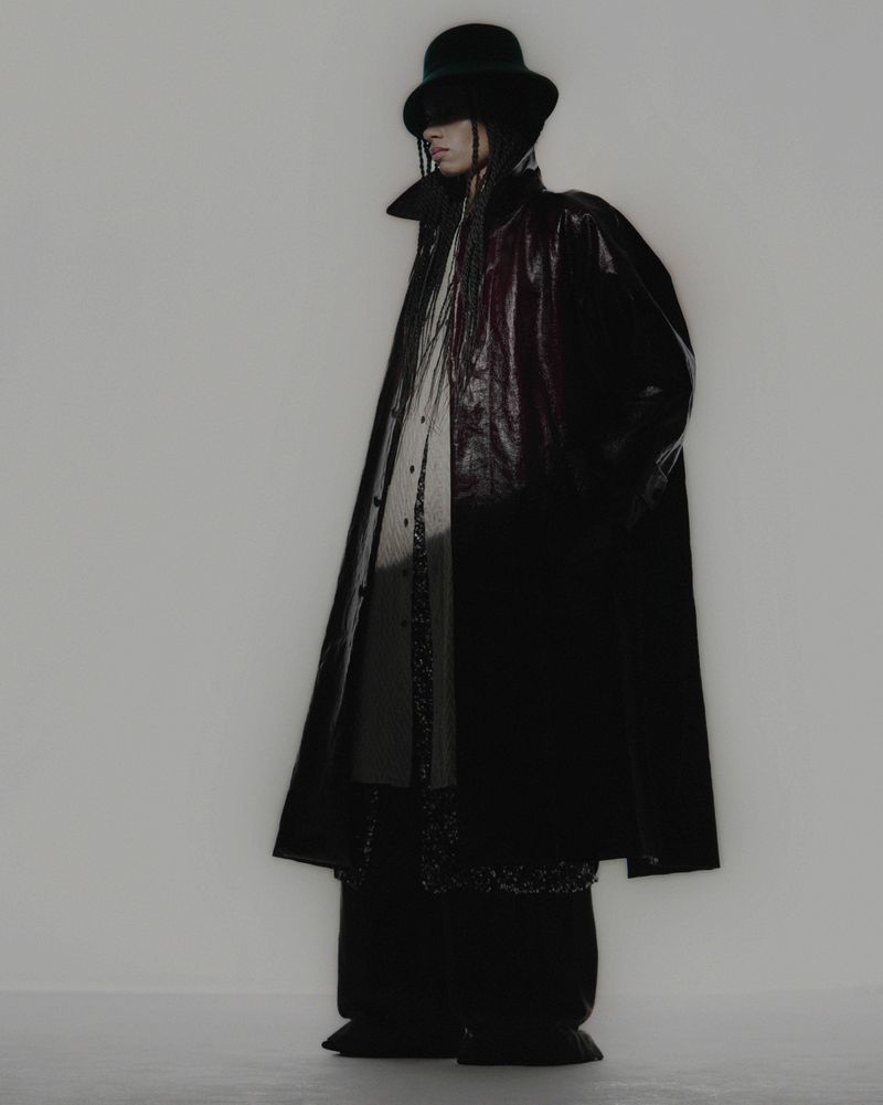 Person in black coat and bowler hat poses in a studio, showcasing dark fashion and style against a plain background.