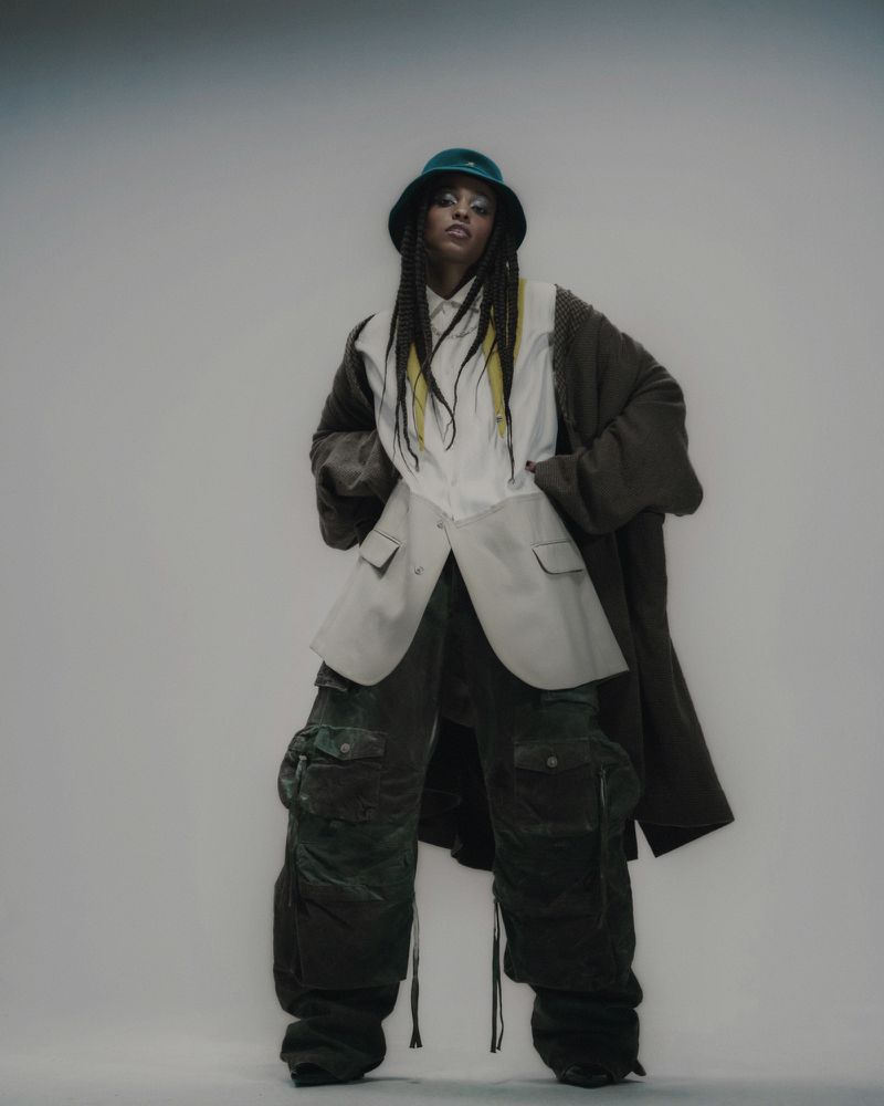 A stylish person in oversized clothing posing in a studio with a bucket hat, long braided hair with yellow streaks, baggy pants and an overcoat.