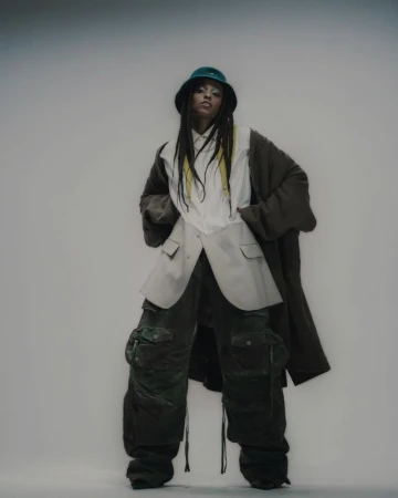 A stylish person in oversized clothing posing in a studio with a bucket hat, long braided hair with yellow streaks, baggy pants and an overcoat.