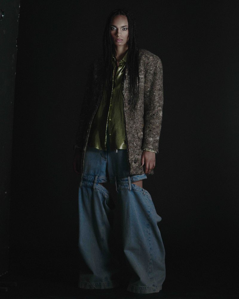 Fashion photo of a Black adult woman in a coat and jeans with cutout legs, posing in a dark indoor setting with soft studio lighting.