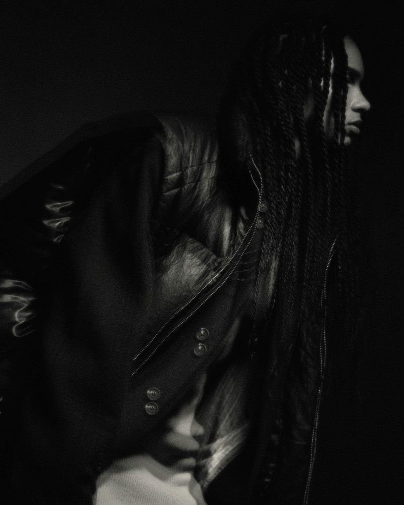 Black and white fashion shot of a woman with long braids wearing a quilted leather jacket in a dimly lit indoor environment.