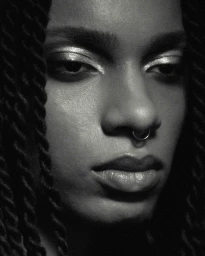 Close-up monochrome portrait of a Black woman with braided hair and a septum ring wearing shimmering eye makeup.