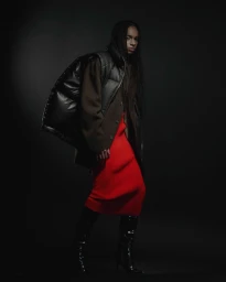 Model with braided hair wearing a glossy black puffer vest over a brown blazer, vibrant red skirt, and black boots against a dark background.