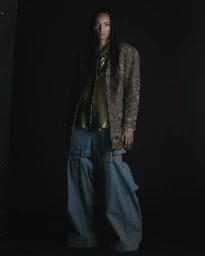 Model with long braided hair wearing a textured coat, olive shirt, and baggy denim pants against a dark background.