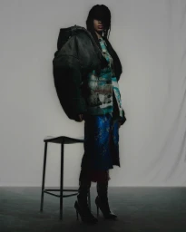 Model wearing a dark oversized puffer jacket and blue patterned skirt standing next to a metal stool