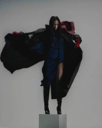 Black woman in a dramatic dark cloak and blue dress standing on a grey pedestal against a neutral studio backdrop.