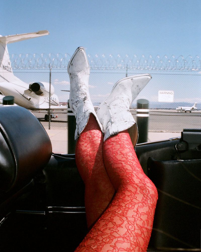 Legs in red lace stockings and white cowboy boots rest on a car door, against a backdrop of a sunny airfield and private jets.