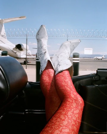 Legs in red lace stockings and white cowboy boots rest on a car door, against a backdrop of a sunny airfield and private jets.