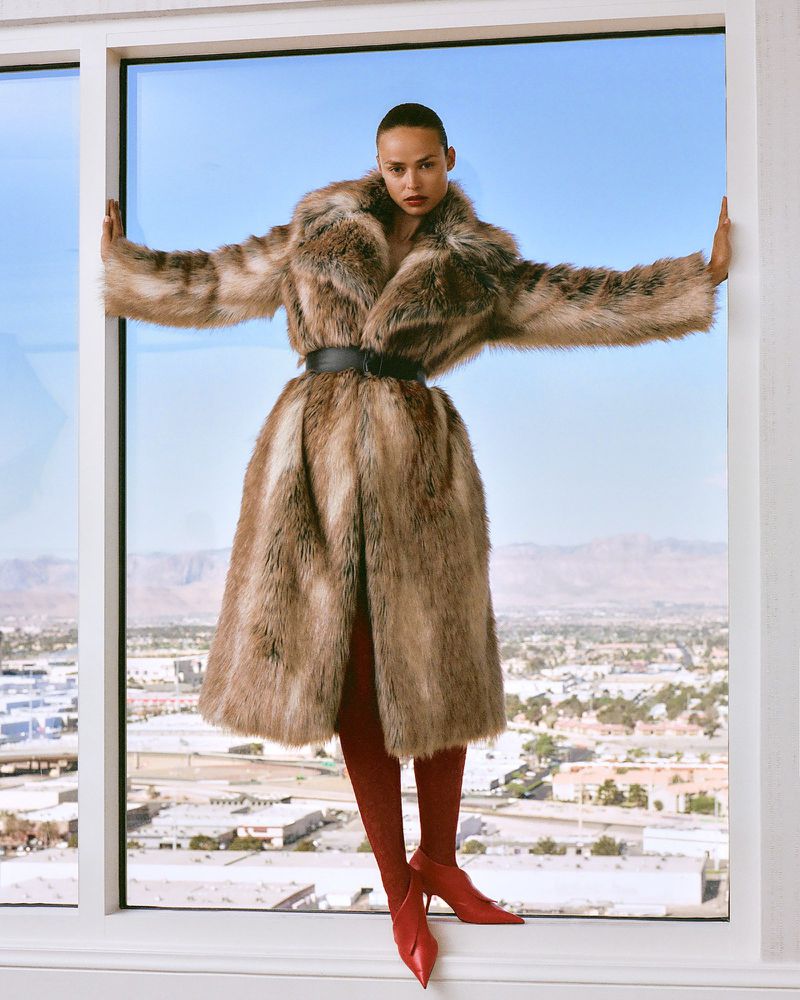 Fashionable woman poses in a fur coat and red tights, standing in front of a high-rise window with city view in the background.