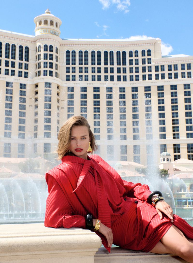 Elegant woman in a red suit poses in front of the Bellagio hotel and fountain in Las Vegas under a blue sky with soft white clouds.