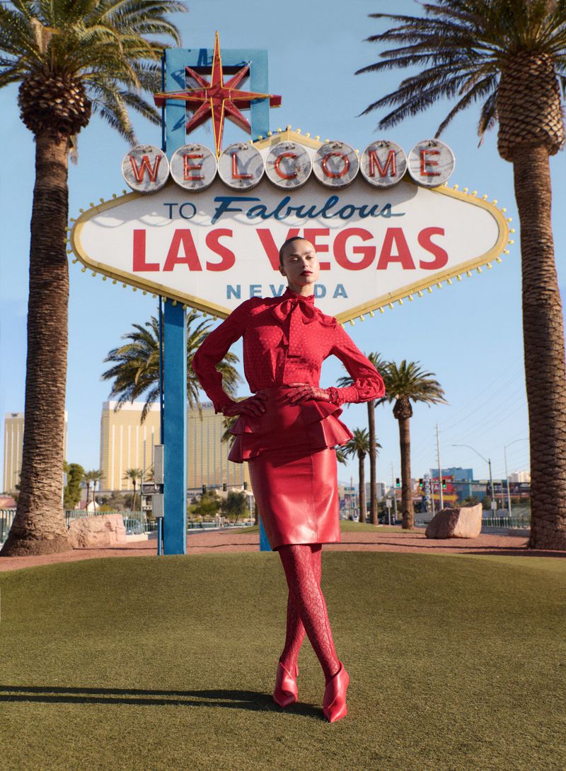 A fashion model in a red outfit poses in front of the Welcome to Fabulous Las Vegas sign on a sunny day with palm trees.