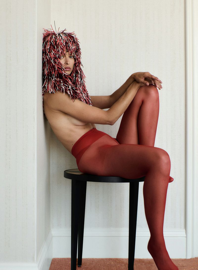Adult female in red tights and an elaborate red-white headdress, seated on a black stool against a neutral wall in a photo studio.