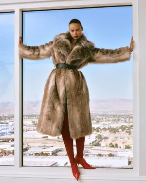 Birgit Kos wearing a fur coat and red boots inside a high-rise window framing a wide city skyline