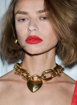 Close-up beauty portrait of model Birgit Kos wearing a bold gold heart necklace and vibrant red lipstick.