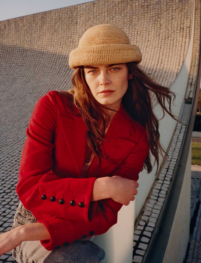 Attractive woman in a red jacket and hat poses outdoors with a neutral expression against a stone and white architectural structure.