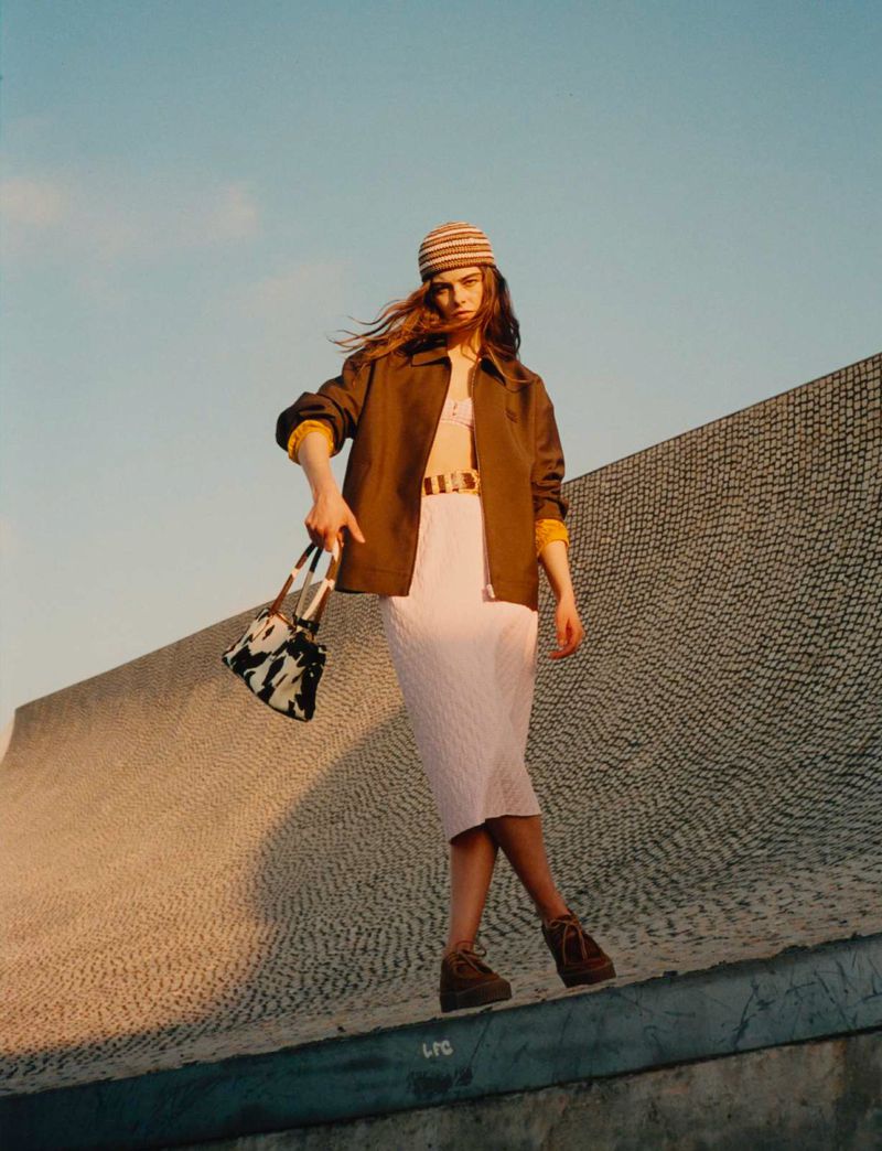 Fashionable woman in jacket, knit skirt and hat poses on a concrete structure with a cow-print handbag in an outdoor environment.
