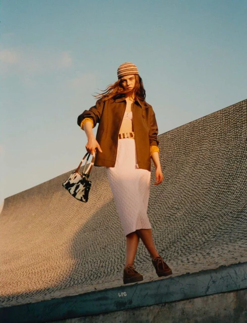 Fashionable woman in jacket, knit skirt and hat poses on a concrete structure with a cow-print handbag in an outdoor environment.