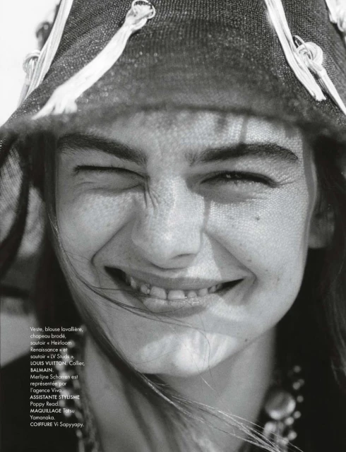 Close-up portrait of a smiling woman wearing a hat with tassels, black and white fashion editorial style shot for a magazine.